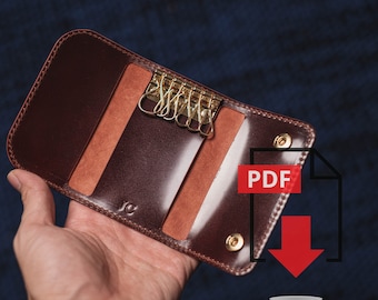 May include: A brown leather key holder with a polished finish, featuring gold-colored key hooks. The key holder is open, revealing the interior and the keys. The initials "JC" are subtly embossed on the leather.