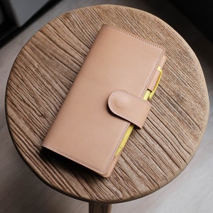 May include: A tan leather planner with a yellow pen tucked inside. The planner has a flap closure with a button.