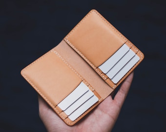 May include: A tan leather card holder featuring a bifold design with visible white stitching along the edges. The interior contains multiple slots, each holding a plain white rectangular card, providing organized storage for several cards.