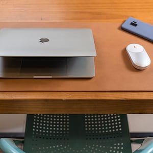 May include: A brown leather desk mat with a closed laptop computer, a white wireless mouse, and a blue smartphone on top.