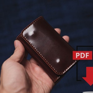 Pu&ograve; includere: Un portachiavi in pelle marrone scuro lucido con chiusura a scatto. Il portachiavi rettangolare &egrave; tenuto in mano su uno sfondo blu scuro. Un'icona PDF rossa con una freccia rivolta verso il basso si trova nell'angolo in basso a destra.