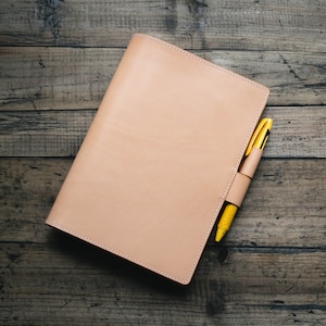 May include: A light tan leather notebook cover with visible stitching along the edges. A bright yellow pen is inserted into a matching leather pen loop attached to the side of the cover. The notebook is rectangular and features a smooth, uniform surface.