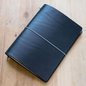 Könnte beinhalten: Ein schwarzes, ledergebundenes Notizbuch mit einem grauen Gummibandverschluss. Das Notizbuch ist geschlossen und liegt flach auf einer Holzoberfläche.