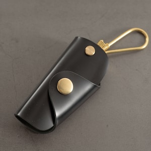 Pode incluir: Um porta-chaves de couro preto com um gancho de metal dourado e dois botões de pressão de metal dourado.