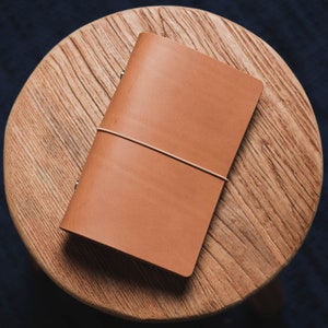 Peut inclure: Un carnet relié en cuir marron avec une fermeture élastique blanche. Le carnet est fermé sur une surface en bois.