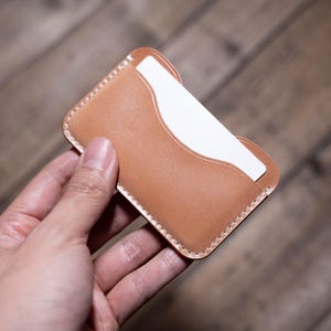 May include: A tan leather card holder with a white card partially visible. The card holder has a stitched border and a curved opening. The item is held in a hand.