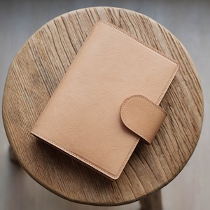 Può includere: Un diario rilegato in pelle color beige con chiusura a lembo su una superficie in legno.