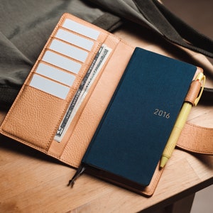 May include: A tan leather wallet with a blue notebook and a yellow pen inside. The wallet has multiple card slots and a billfold. The notebook has the year 2016 printed on the cover.