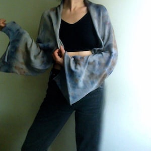 Peut inclure: Un châle à motif tie-dye avec de larges manches évasées. Le châle présente des nuances de bleu et de marron. La personne sur l'image porte un haut noir et un jean foncé.
