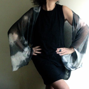 Peut inclure: Une robe noire sans manches avec un cardigan transparent à motifs gris et blancs. Le cardigan est ample, fluide et ouvert sur le devant.