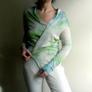 Peut inclure: Une femme portant une chemise à manches longues en tie-dye vert et bleu sur fond blanc. La chemise est enroulée autour de son corps et nouée à la taille. Elle porte un pantalon blanc.