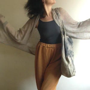 Peut inclure: Une femme porte un cardigan beige clair à manches longues et larges, un débardeur noir et un pantalon moutarde. Un grand sac fourre-tout avec un motif bleu-gris est porté sur l'épaule.