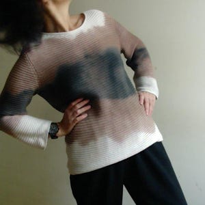 May include: A long-sleeved sweater with a ribbed texture, featuring a color gradient from white to brown and black. The sweater has a crew neck and flared sleeves. The person is wearing black pants.