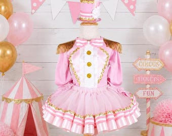 May include: A pink circus ringmaster costume featuring a long-sleeve jacket with gold fringe epaulets, a white ruffled front with gold buttons, and a layered tulle skirt. The outfit includes a matching pink top hat with a white feather plume.