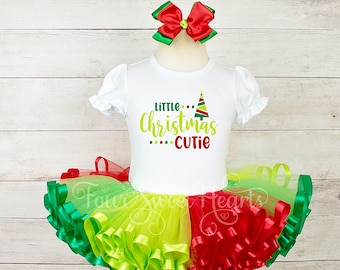 May include: A white Christmas-themed tutu with green, yellow, and red ribbon. The tutu is paired with a white t-shirt that says "Little Christmas Cutie" with a green, red, and yellow Christmas tree graphic. A red bow with green leaves is on top of the tutu.
