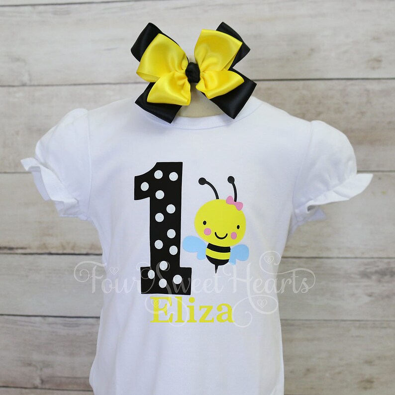 May include: White short-sleeved shirt with a black and white polka-dotted number 1 and a cartoon bee graphic. The shirt has the name "Eliza" in yellow script. A large yellow and black bow is attached to the top of the shirt.