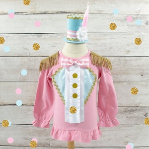 May include: A pink long-sleeved shirt with gold fringe epaulets and a decorative bib. The bib has a light blue heart shape with gold trim and three gold buttons. A matching light blue and pink top hat with a feather sits above the shirt.