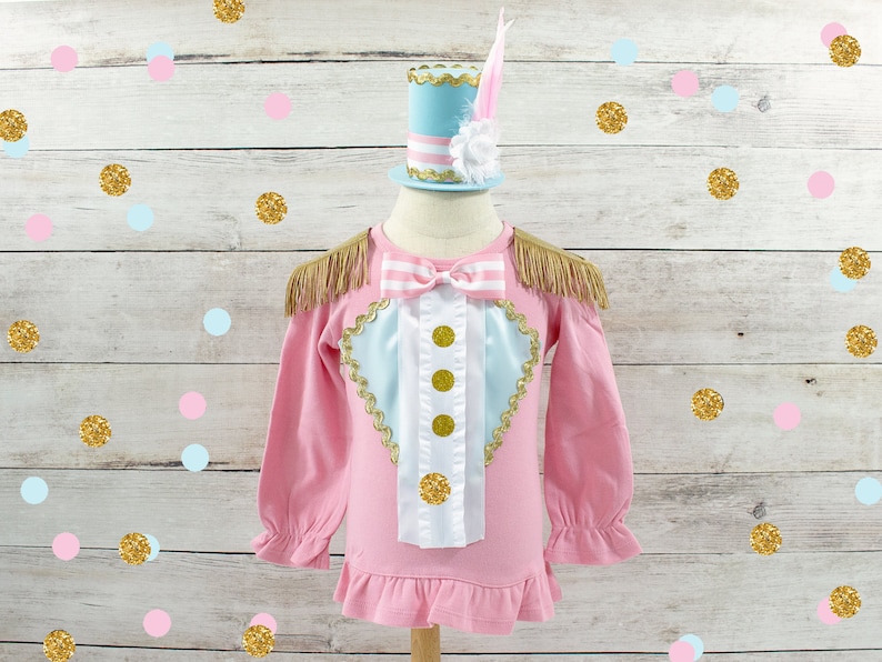May include: A pink long-sleeved shirt with gold fringe epaulets and a decorative bib. The bib has a light blue heart shape with gold trim and three gold buttons. A matching light blue and pink top hat with a feather sits above the shirt.