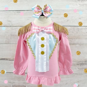 May include: Pink long-sleeved shirt with a circus-themed design. The shirt features a light blue and white heart-shaped design with gold trim and three gold buttons. Gold fringe accents the shoulders. A matching bow is at the top.