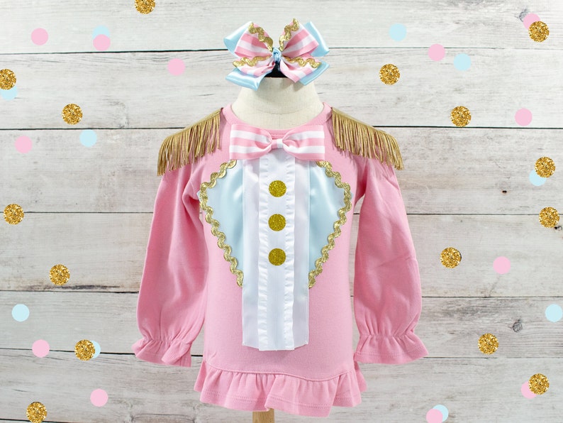 May include: Pink long-sleeved shirt with a circus-themed design. The shirt features a light blue and white heart-shaped design with gold trim and three gold buttons. Gold fringe accents the shoulders. A matching bow is at the top.