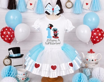 Puede incluir: Una camiseta blanca de manga corta y una falda de tul azul y blanca con capas. La camiseta presenta un gráfico de un sombrero de copa, tazas apiladas, el número 1 y el texto Lilliana In ONEderland. Incluye un lazo para el pelo a juego.