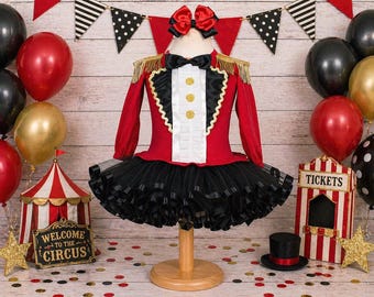 May include: A red and black circus-themed dress featuring a white ruffled front panel with three gold buttons, a black bow tie, gold fringe shoulder epaulets, and a voluminous black tulle skirt. A matching red and black hair bow is included.