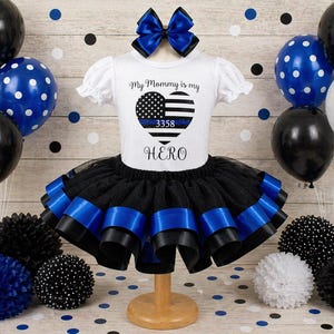 May include: A white short-sleeved outfit with a black tutu skirt and blue ribbon trim. The shirt has the text "My Mommy is my HERO" with a heart design. A blue bow and balloons complete the look.