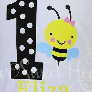 May include: White t-shirt featuring a large black number one with white polka dots, and a cartoon bee with a pink bow. The name "Eliza" is printed in yellow below the number and bee.