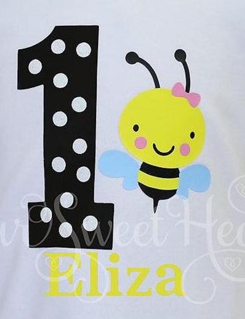 May include: White t-shirt featuring a large black number one with white polka dots, and a cartoon bee with a pink bow. The name "Eliza" is printed in yellow below the number and bee.