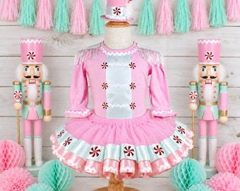May include: A pink and light blue long-sleeve dress with a tutu skirt, featuring a matching top hat, silver fringe shoulder accents, and peppermint candy graphics on the bodice and skirt hem, displayed on a mannequin between two nutcrackers.