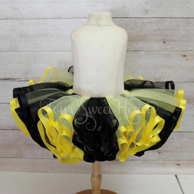 May include: A yellow, black, and white tutu skirt displayed on a mannequin. The skirt features layers of tulle and yellow satin ribbon trim. The text "Our Sweet Heart" is visible on the skirt.