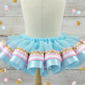 May include: A light blue tutu skirt with layers of tulle and decorative ribbons. The ribbons are light blue with pink and white stripes, and gold trim. The skirt is displayed on a mannequin.