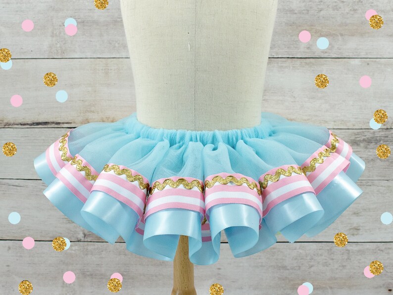 May include: A light blue tutu skirt with layers of tulle and decorative ribbons. The ribbons are light blue with pink and white stripes, and gold trim. The skirt is displayed on a mannequin.