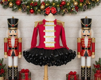 May include: A red long-sleeve dress with gold fringe epaulets, a white central panel with gold trim, and a black tulle skirt. A red bow is attached at the neckline. The dress is displayed on a mannequin between two wooden nutcracker figures.