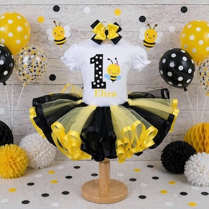 May include: A white short-sleeved shirt with a bee graphic and the number 1, paired with a black and yellow tutu. The ensemble is decorated with balloons, paper honeycomb balls, and bee cutouts, creating a festive birthday theme.