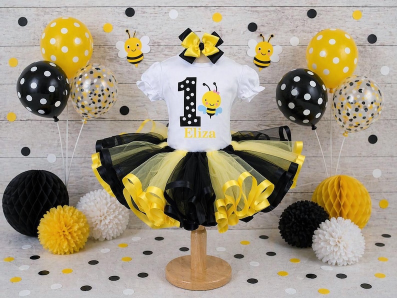 May include: A white short-sleeved shirt with a bee graphic and the number 1, paired with a black and yellow tutu. The ensemble is decorated with balloons, paper honeycomb balls, and bee cutouts, creating a festive birthday theme.