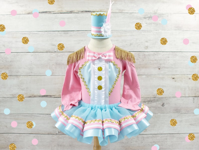May include: A pink and blue circus-themed outfit with a matching hat. The long-sleeved top has gold fringe epaulets and gold button details. The tutu skirt has layers of blue tulle and pink and white ribbon trim. The hat is light blue with a white feather.