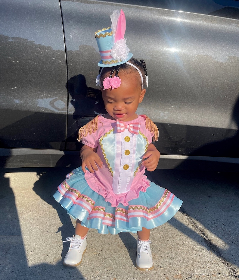 May include: A child wearing a pink and blue circus-themed outfit with a matching top hat. The outfit features gold trim, a bow tie, and a tutu-style skirt. The child is also wearing white shoes and a white headband with pink flowers.
