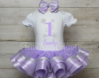 May include: A white short-sleeved bodysuit with a lavender number one and snowflake graphics, paired with a layered lavender tulle tutu skirt featuring silver ribbon trim. A matching lavender hair bow with silver accents is included.