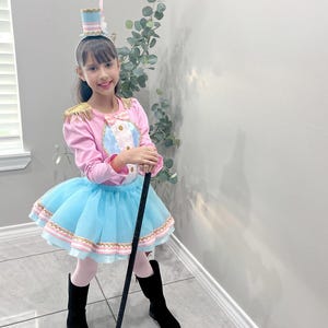 May include: A child's circus ringmaster costume. The outfit includes a pink long-sleeved top with gold epaulets and a blue and white center, a light blue tutu skirt, black boots, and a small top hat. A black cane is held.