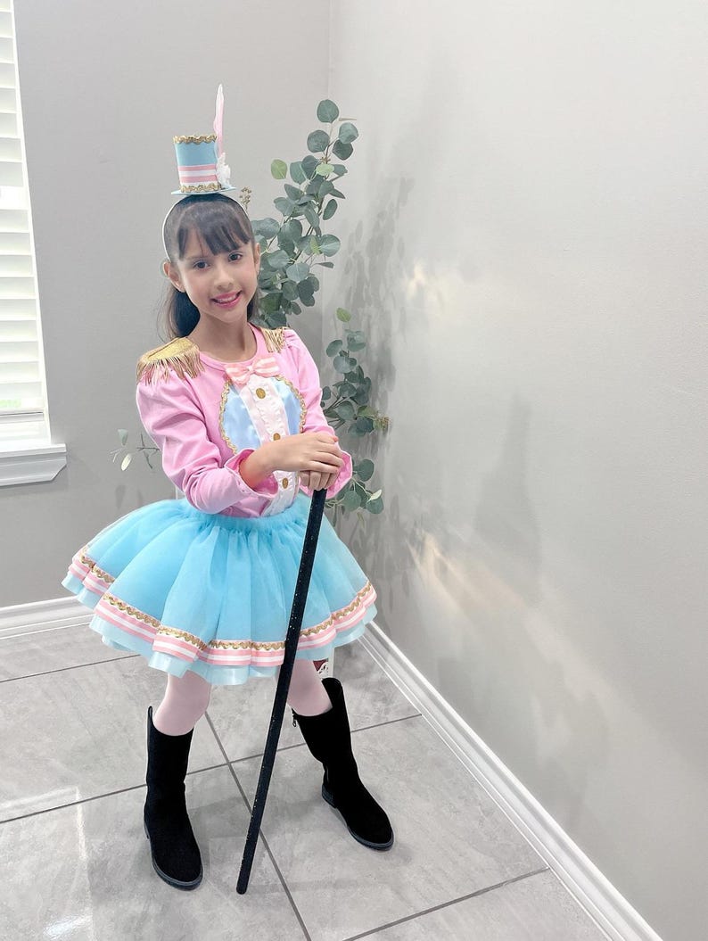 May include: A child's circus ringmaster costume. The outfit includes a pink long-sleeved top with gold epaulets and a blue and white center, a light blue tutu skirt, black boots, and a small top hat. A black cane is held.