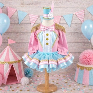 May include: A pink and blue circus-themed outfit with gold accents, a matching hat, and a small circus tent. Balloons in pink and blue are in the background, along with a decorative stand and pennant banner. The outfit is designed for a child.