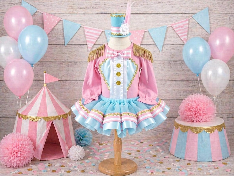 May include: A pink and blue circus-themed outfit with gold accents, a matching hat, and a small circus tent. Balloons in pink and blue are in the background, along with a decorative stand and pennant banner. The outfit is designed for a child.