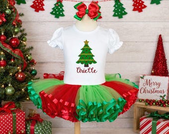 May include: A white Christmas-themed outfit with a green and red tutu skirt, a matching bow, and a shirt with a Christmas tree graphic and the name "Arielle". Festive decorations include a Christmas tree and a "Merry Christmas" sign.