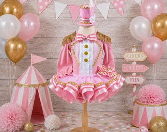 May include: A pink circus-themed costume featuring a long-sleeved jacket with gold fringe epaulets, a white front panel with three gold buttons, and a layered tutu skirt. The outfit includes a matching pink top hat with a white feather accent.
