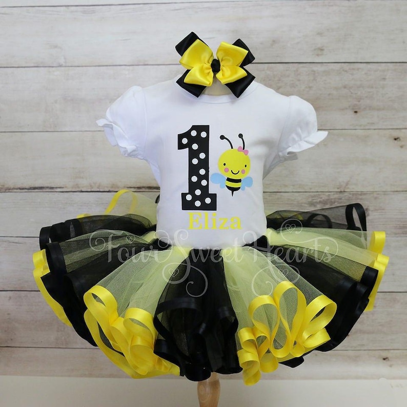 May include: A white short-sleeved shirt with a bee graphic and the number 1. The shirt is paired with a black and yellow tutu skirt and a matching bow. The name "Eliza" is printed on the shirt.