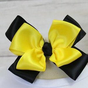 May include: A large hair bow featuring a layered design with black and yellow satin ribbons. The bow is centered with a small black knot and is likely attached to a hair clip or band. The bow is set against a white background.