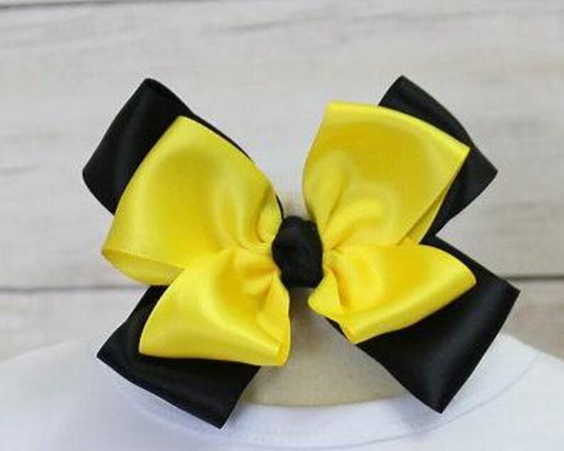 May include: A large hair bow featuring a layered design with black and yellow satin ribbons. The bow is centered with a small black knot and is likely attached to a hair clip or band. The bow is set against a white background.