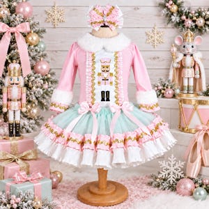 May include: A long-sleeved dress featuring a pink bodice with a central nutcracker graphic, a white faux fur collar, and a layered tulle skirt in mint green and pink with gold trim. The dress includes two pink bows and a matching hair bow.