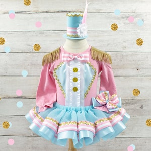 May include: A pink and blue circus-themed outfit with gold accents. The long-sleeved top has gold fringe epaulets and gold buttons. The tutu skirt is light blue with pink and white ribbon trim. A matching hat with a feather is included.